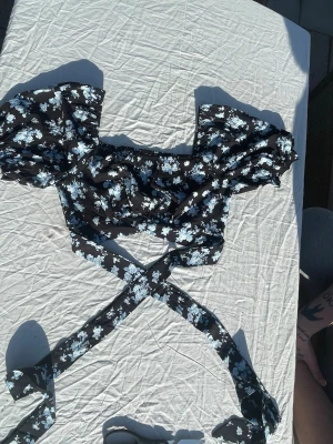 Blommig svart croppad blus med knytning - Säljer en söt croppad blus som är svart och har vita blommor. Den har puffiga korta ärmar och långa knytband som man knyter runt midjan för en trendig look. Endast testad och säljes pga för liten🤩☺️