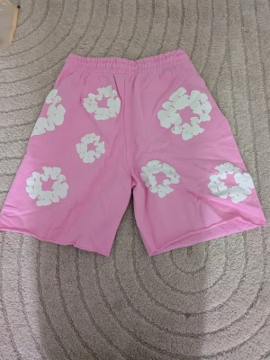 Rosa shorts med vita blommor  - Säljer ett par rosa shorts med elastisk midja och snörning. Shortsen har stora vita blommönster över hela plagget och är tillverkade i ett mjukt, bekvämt material. Perfekta för varma dagar och en avslappnad stil.