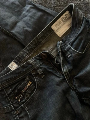 Mörkgrå jeans med dragkedjedetalj - Snygga mörkgrå/blå jeans från South East G-STAR RAW med raka ben och cool dragkedjedetalj vid fickan + coola ”smådetaljer”. Lite oklart vilken storlek men skulle gissa på XS/S. 