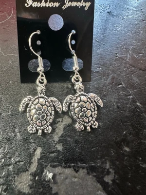 Silvriga örhängen med sköldpaddor - Unika silvriga örhängen med detaljerade sköldpaddor i metall. Hängande modell med krok, perfekt för dig som gillar djurmotiv och vill sticka ut lite extra. Säljs på originalkort.