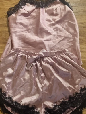 Rosa satinset med spetsdetaljer - Sött tvådelat set i ljusrosa satin med svarta spetsdetaljer. Linnet är ärmlöst och har spets längs urringningen, medan shortsen har en rosett i midjan och spets längs bensluten. Perfekt för en lyxig känsla hemma