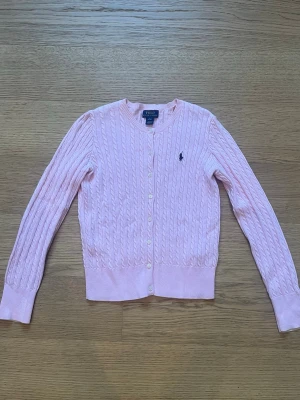 Ljusrosa kabelstickad kofta Polo Ralph Lauren - Supersöt ljusrosa kabelstickad kofta från Polo Ralph Lauren med rund halsringning och knappar framtill. Klassisk marinblå logga broderad på bröstet och ribbade muddar vid ärmslut och nederkant. Perfekt för lager på lager och en preppy look.