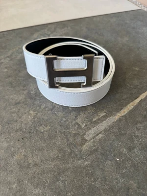 Vitt skärp med metallspänne - Snyggt vitt skärp med ett stort, silvrigt metallspänne i form av ett H. Skärpet har en stilren och modern look, perfekt för att lyfta vilken outfit som helst. Tillverkat i slätt skinn med fina sömmar längs kanterna.