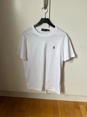 Vit t-shirt från Polo Ralph Lauren - Klassisk vit t-shirt från Polo Ralph Lauren med korta ärmar och rund hals. T-shirten har en liten broderad logga på bröstet och är tillverkad i mjuk bomull. Perfekt basplagg för en clean och stilren look.