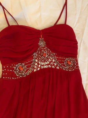 Röd långklänning med strassdetaljer - Elegant röd långklänning med tunna axelband och vackra strass- och pärldetaljer över bysten. Klänningen har en lätt rynkad överdel och ett flytande, svepande kjolparti. Perfekt för bal eller festliga tillfällen.