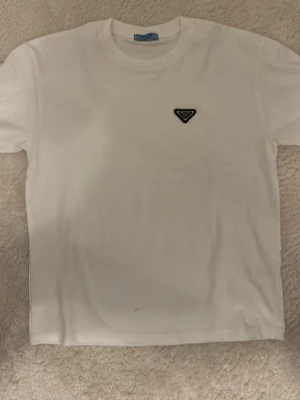 Vit t-shirt från Prada med logga - Snygg vit t-shirt från Prada med klassisk rund hals och korta ärmar. På bröstet sitter Pradas ikoniska trekantslogga i svart. Perfekt basic-plagg med exklusiv känsla och stilren design. Tillverkad i mjuk bomull för skön komfort.