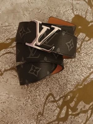 L V svart monogram bälte - Snyggt svart bälte från L V med klassiskt monogrammönster och stor L V-spänne i silver. Bältet är gjort i skinn och har en exklusiv känsla som lyfter vilken outfit som helst.