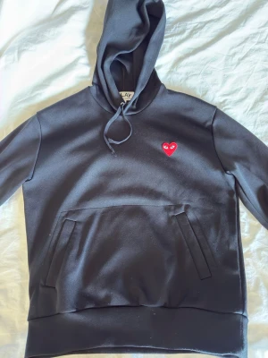 Svart hoodie från Comme des Garçons Play - Svart hoodie med huva och dragsko från Comme des Garçons Play. På bröstet sitter det ikoniska röda hjärtat med ögon. Hoodien har en stor magficka och är tillverkad i mjukt material som känns skönt mot huden. Nypris:2,3 och självklart äkta