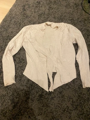 Beige öppen kofta i tunt material - Säljer en beige, långärmad öppen kofta i tunt och mjukt material. Koftan har en avslappnad passform utan knappar och faller snyggt framtill. Perfekt att slänga över en topp för en enkel och stilren look.