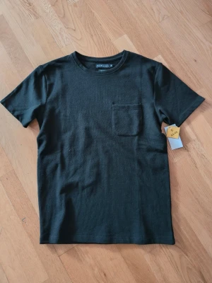 Svart t-shirt med bröstficka  - En stilren svart t-shirt  med rund halsringning och en enkel bröstficka på vänster sida. Tillverkad i mjuk bomull och har en klassisk passform med korta ärmar. Perfekt basplagg för alla tillfällen.