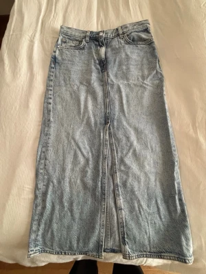 Lång jeanskjol i ljusblå denim från Arket - Trendig lång jeanskjol i ljusblå denim med klassiska femficksdetaljer och slits framtill. Perfekt för dig som gillar en avslappnad och cool stil. Kjolen har knapp- och dragkedjestängning samt bälteshällor. Nypris ca790kr