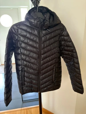 Svart dunjacka med huva - Snygg svart dunjacka med huva och dragkedja framtill. Jacka har ett quiltat mönster i diagonala linjer och två fickor med dragkedja. Perfekt för kyliga dagar och enkel att matcha med olika outfits.