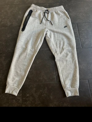 Gråa mjukisbyxor från Nike - Säljer ett par gråa mjukisbyxor från Nike med svart logga på vänster ben. Byxorna har resår och snörning i midjan, muddar vid bensluten och två sidofickor. Perfekta för chill dagar eller träning. Klassisk och enkel design i mjukt material. Känns som M