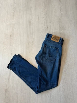 Blå slim fit jeans från Replay - Säljer ett par blå slim fit jeans från Replay med klassisk femficksdesign och snygga slitningar framtill. Jeansen har en skinnpatch med logga bak i midjan och är tillverkade i mjukt denimtyg för en bekväm känsla.
