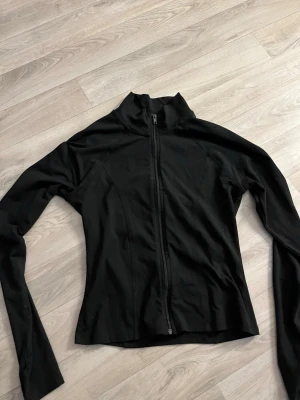 Svart sportig jacka med dragkedja - Svart, figursydd jacka med hög krage och hel dragkedja framtill. Jackan har lång ärm och är tillverkad i ett stretchigt material som passar perfekt för en aktiv livsstil. Det är hål för tummarna på ärmarna. Katt finns i hemmet