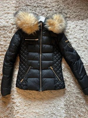 Svart pufferjacka Rockandblue - Snygg svart pufferjacka med stor luva kantad av beige fuskpäls. Jackan har guldfärgade dragkedjor, quiltade detaljer på sidorna och ärmarna samt en patch på ena ärmen. Perfekt för kalla vinterdagar och riktigt trendig look.