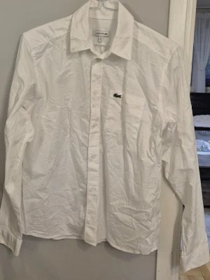 Vit skjorta från Lacoste - Klassisk vit skjorta från Lacoste med lång ärm och knappar framtill. Skjortan har en bröstficka med den ikoniska gröna krokodilloggan. Perfekt till både jeans och chinos.