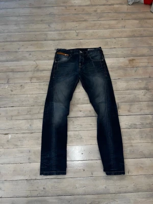 Mörkblå jeans från Jack & Jones - Snygga mörkblå jeans från Jack & Jones med klassisk femficksdesign och diskreta slitningar. Jeansen har raka ben och orangea detaljer vid fickorna. Perfekta för en trendig och avslappnad look. Storlek 32:34 hör sv dig vid minsta fundering