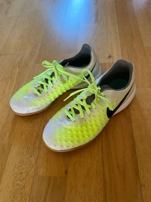 Nike vita och neongula fotbollsskor - Säljer ett par Nike fotbollsskor i vitt och neongult med svart swoosh på sidan. Skorna har snörning och en texturerad ovansida för bättre bollkontroll. Inomhusskor. Vet inte vilken storlek skorna är i men passar mig som har storlek 38.