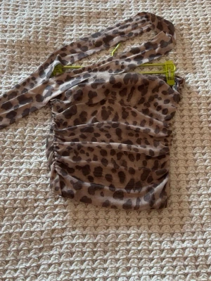 Leopardmönstrad one shoulder topp - Trendig one shoulder topp med leopardmönster i bruna och beige toner. Toppens material är lätt och stretchigt, med rynkade detaljer längs sidorna för en snygg passform. Perfekt för dig som vill sticka ut med en djurisk look.