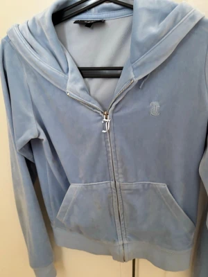 Blå velour hoodie från Juicy Couture - Säljer en ljusblå velour hoodie från Juicy Couture med dragkedja framtill och broderad logga på bröstet. Hoodien har stora fickor och en klassisk huva. Perfekt för en chill och trendig look.