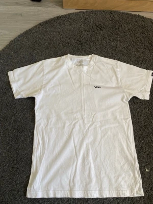 Vit t-shirt från Vans - Klassisk vit t-shirt från Vans med liten logga på bröstet. T-shirten har rund halsringning och korta ärmar, perfekt för en avslappnad och stilren look. Tillverkad i mjuk bomull som känns skön mot huden.