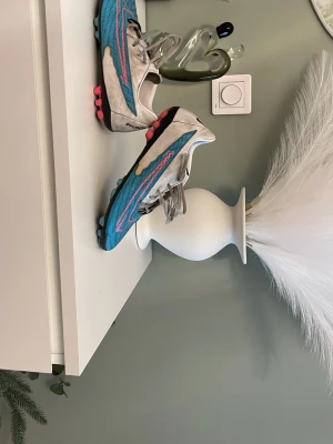 Nike blå och vita fotbollsskor - Säljer ett par Nike fotbollsskor med blå ovandel, vita sidor och rosa detaljer. Skorna har snörning och dobbar för spel på gräs. Swoosh-loggan syns tydligt på sidan. Perfekta för dig som vill sticka ut på planen.