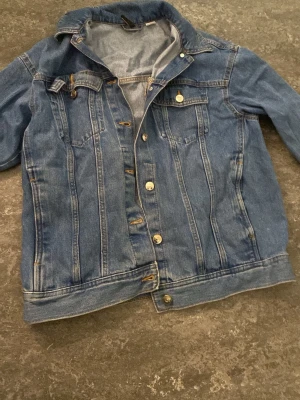 Klassisk blå jeansjacka - Snygg blå jeansjacka med raka sömmar och klassiska bröstfickor med lock. Jackan har silverfärgade knappar framtill och en tidlös design som passar till det mesta. Perfekt för dig som gillar en avslappnad och trendig stil.