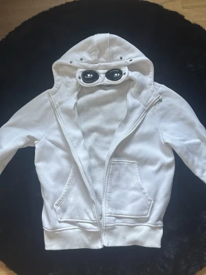 Vit hoodie med glasögon från C.P. Company - Säljer en vit hoodie från C.P. Company med dragkedja och ikoniska glasögon i huvan. Hoodien har två fickor framtill och ribbade muddar. Perfekt för dig som vill sticka ut med en unik streetwear-stil.