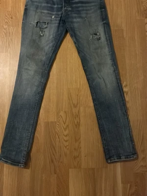 Jack & Jones jeans  - Säljer ett par blå s jeans med slitningar och hål på båda benen framtill. Jeansen har klassisk femficksdesign och är i en tvättad denim med snyggt sliten look. Perfekta för dig som gillar en trendig och avslappnad stil.