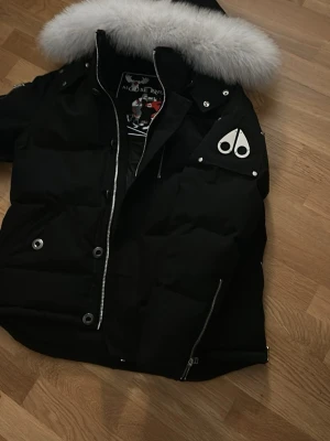 Svart dunjacka Moose Knuckles päls - Svart dunjacka från Moose Knuckles med stor vit fuskpäls på huvan och ikoniska loggan på axeln. Jackan har silvriga dragkedjor, tryckknappar och detaljer, samt quiltad design. Perfekt för kalla vinterdagar och riktigt snygg streetstil.