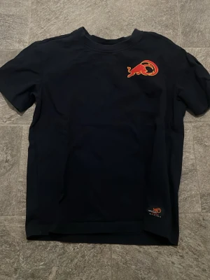 Svart Red Bull Racing t-shirt - Snygg svart t-shirt från Red Bull Racing med deras ikoniska röda tjur-logga på bröstet och en mindre logga vid nederkanten. Klassisk passform och rund halsringning. Perfekt för dig som gillar motorsport och streetwear.