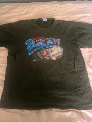 Vintage T-shirt - Grön t-shirt från Bulletin Athletic med stort Toronto Blue Jays-tryck på bröstet. Klassisk rund hals och korta ärmar. Trycket har blå, röd och vit färg med basebollmotiv. Perfekt för dig som gillar sportiga och retro plagg. Pris kan diskuteras. Skriv för mer bilder!