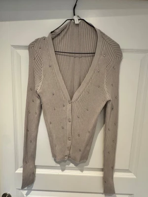 Beige ribbad kofta med pärlor H&M - Snygg beige ribbad kofta från H&M med v-ringning och knäppning framtill. Koftan har långa ärmar och är dekorerad med små pärlor över hela plagget för en extra touch. Perfekt att slänga över en topp eller klänning.