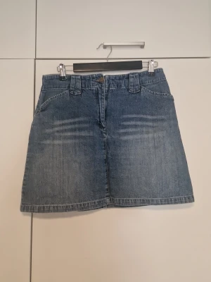 Blå jeanskjol med fickor - Snygg blå jeanskjol i klassisk modell med stora bälteshällor och fickor fram. Kjolen har en lätt A-linjeform och stängs med knapp och dragkedja. Perfekt till sneakers eller boots för en cool vardagslook.