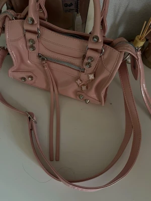 Ljusrosa axelremsväska med nitar - Söt ljusrosa handväska i glansigt material med silverfärgade nitar och dragkedjor. Väskan har både handtag och justerbar axelrem. Perfekt för dig som gillar en trendig och feminin look.
