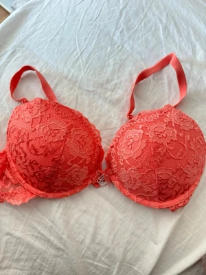 Korallrosa push-up bh med spets - Snygg korallrosa push-up bh med vadderade kupor och vacker blommig spets över hela ytan. Justerbara axelband och liten dekorativ detalj mellan kuporna. Perfekt för dig som gillar färg och spets.