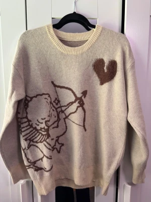 Beige stickad tröja - Stickad beige tröja från BRMG med brunt motiv av en amor med pilbåge och hjärta framtill. På ryggen står texten 'I WOULD RATHER NOT LOVE' i brunt. Tröjan har rund halsringning och ribbade muddar vid ärmslut och nederkant.