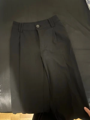 Svarta vida kostymbyxor - Snygga svarta kostymbyxor med vida ben och hög midja. Byxorna har hällor för skärp och stängs med knapp och dragkedja. Perfekta för en stilren look. Materialet ser ut att vara en mjuk och följsam textil.