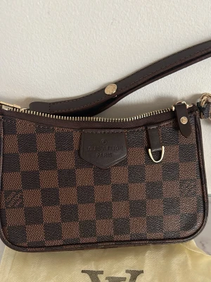 Brun axelväska  - Säljer en klassisk  axelväska i brunt Damier-mönster. Väskan har guldfärgade metalldetaljer, dragkedja och avtagbar handledsrem i mörkbrunt läder. Perfekt storlek för det viktigaste och ikonisk logga framtill. Använd ett par få gånger, den har gått sönder lite vid handtagen men funkar att använda fortfarande 