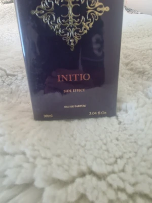 Initio Side Effect Eau de Parfum 90ml - Initio Side Effect Eau de Parfum i en elegant mörklila kartong med gulddetaljer. Flaskan rymmer 90 ml och doften är känd för sin lyxiga och unika karaktär. Förpackningen har exklusiv design med dekorativt guldfärgat mönster.