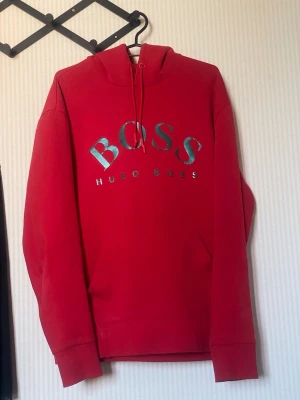 Röd hoodie från Hugo Boss - Snygg röd hoodie från Hugo Boss med stor BOSS-logga i metallic på bröstet. Klassisk modell med huva och dragsko samt magficka framtill. Perfekt för en avslappnad och trendig streetstyle-look.