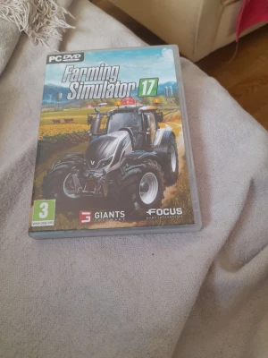 Farming Simulator 17 PC-spel - Farming Simulator 17 till PC, ett realistiskt simuleringsspel där du styr traktorer och maskiner på landsbygden. Omslaget visar en stor svart traktor mot en lantlig bakgrund. Spelet ges ut av GIANTS Software och Focus Home Interactive.