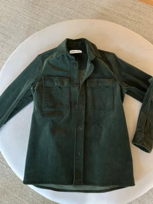 Grön manchester overshirt från Samsoe Samsoe - Stilren mörkgrön overshirt i manchester från Samsoe Samsoe. Skjortan har två stora bröstfickor med lock och knappar, klassisk krage och raka ärmar med knapp vid ärmslut. Perfekt lager-på-lager-plagg med snygg struktur och avslappnad passform.