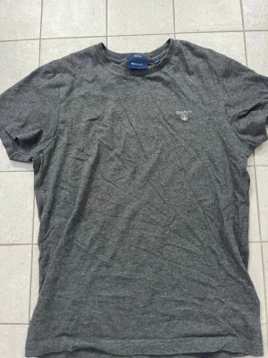 Mörkgrå t-shirt från GANT - Snygg mörkgrå t-shirt från GANT i regular fit med liten broderad logga på bröstet. Klassisk rund halsringning och korta ärmar. Perfekt basplagg i garderoben för en stilren look.