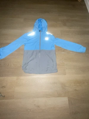 Blå och grå vindjacka från Under Armour - Snygg tvåfärgad vindjacka från Under Armour i ljusblått och grått. Jackan har huva, hel dragkedja framtill och två fickor med dragkedja. Reflekterande detaljer på axlarna för extra synlighet. Perfekt för blåsiga dagar. Som inte säljs längre.