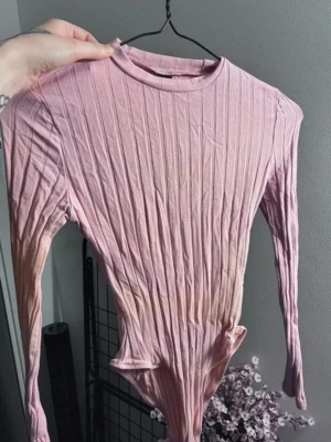 Ribbad rosa body med lång ärm - Säljer en ribbad body i ljusrosa med lång ärm och rund halsringning. Materialet är stretchigt och följsamt, och färgen skiftar lätt mot beige och lila nedtill. Aldrig använd