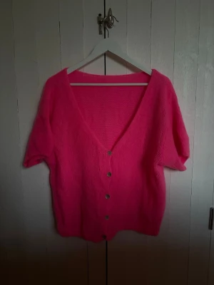 Rosa kofta med kort ärm och knappar - Superfin stickad kofta i starkt rosa med korta ärmar och djup V-ringning. Koftan har silvriga knappar framtill och är gjord i ett mjukt, fluffigt material. Perfekt för att liva upp din outfit!
