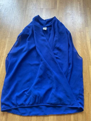 Kornblå ärmlös blus från Vila - Snygg kornblå blus från Vila i storlek M. Blusen är ärmlös med omlott-design framtill och har en lätt, luftig känsla. Fin blus som korsar över bröstet.  Finns även ett ”inbyggt” linne under. Väldigt fint skick, med en liten skavank där det har gått upp lite, se sista bilden. Jag skulle säga att den är mörkblå.  