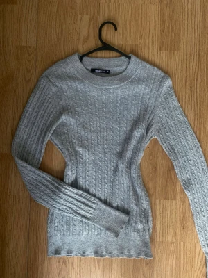 Grå kabelstickad tröja Gina Tricot - Snygg grå kabel stickad tröja från Gina Tricot med rund halsringning och långa ärmar. Tröjan har ett klassiskt kabelmönster och ribbade muddar vid ärmslut och nederkant. Perfekt för lager-på-lager och enkel att matcha med jeans eller kjol. Aldrig använd